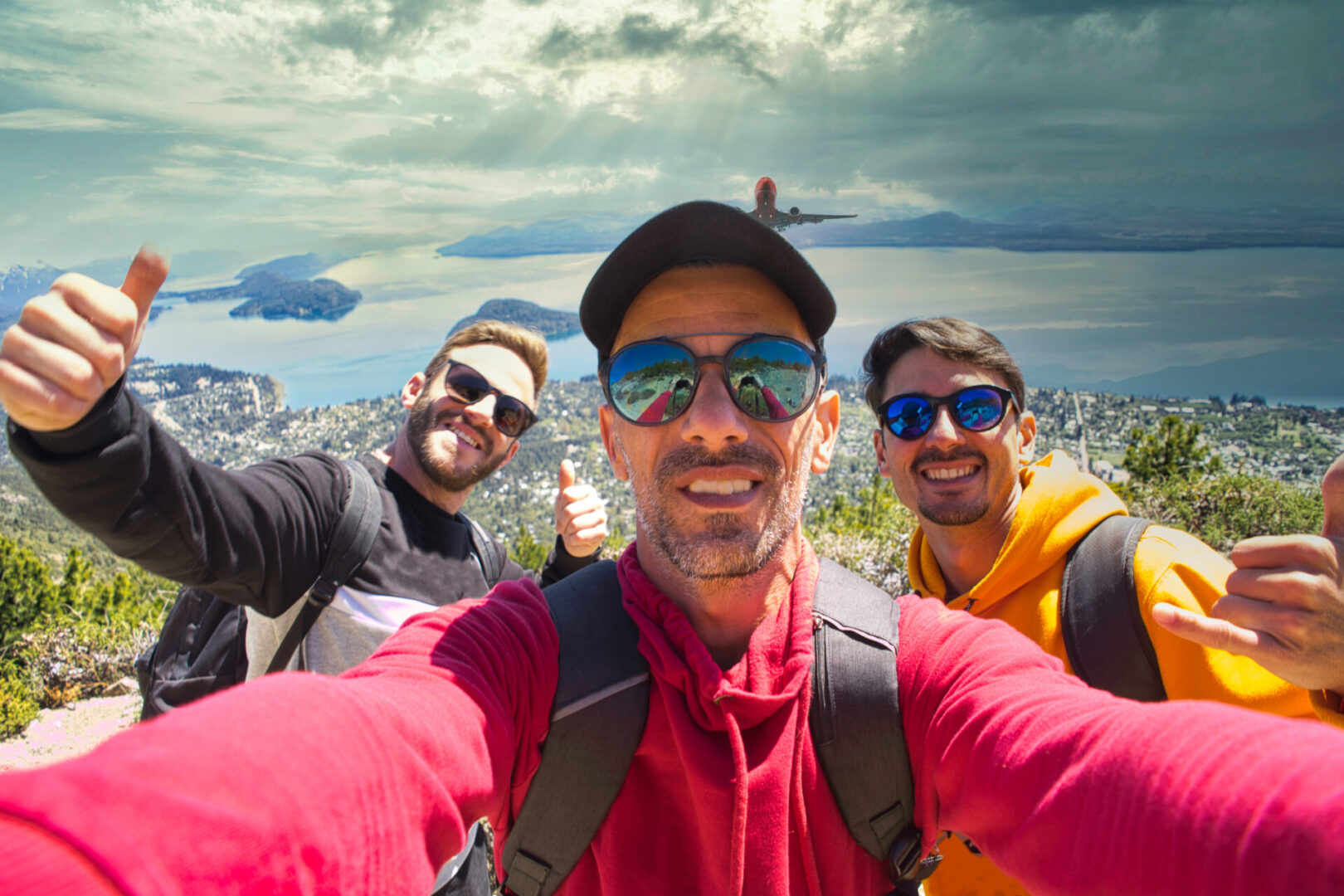 Selfie of three friends with sunglasses and backpack up Cerro Otto in Bariloche with epic view of the city, lake and mountains with cloudy skies and sunrays.